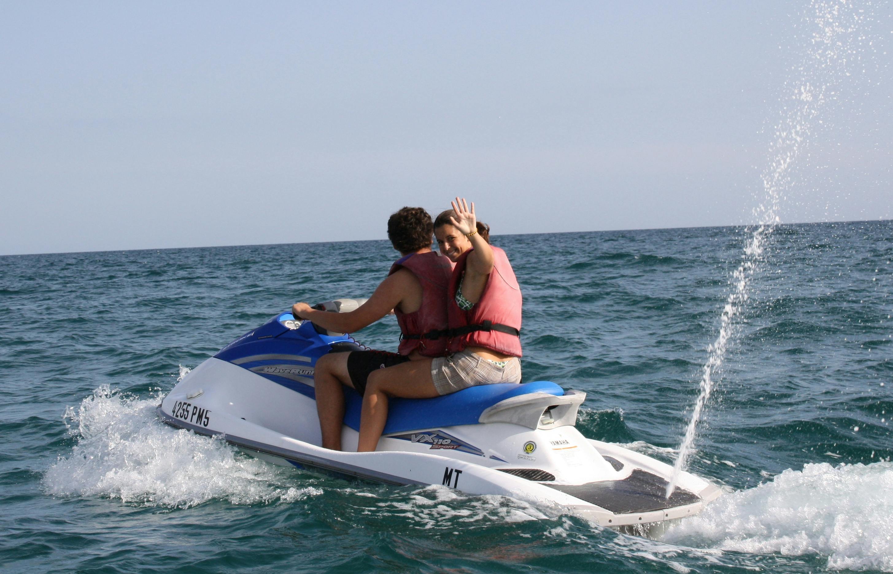 ライフジャケットを着た2人が海上でジェットスキーに乗る。 背中の人がカメラに向かって陽気に手を振る。