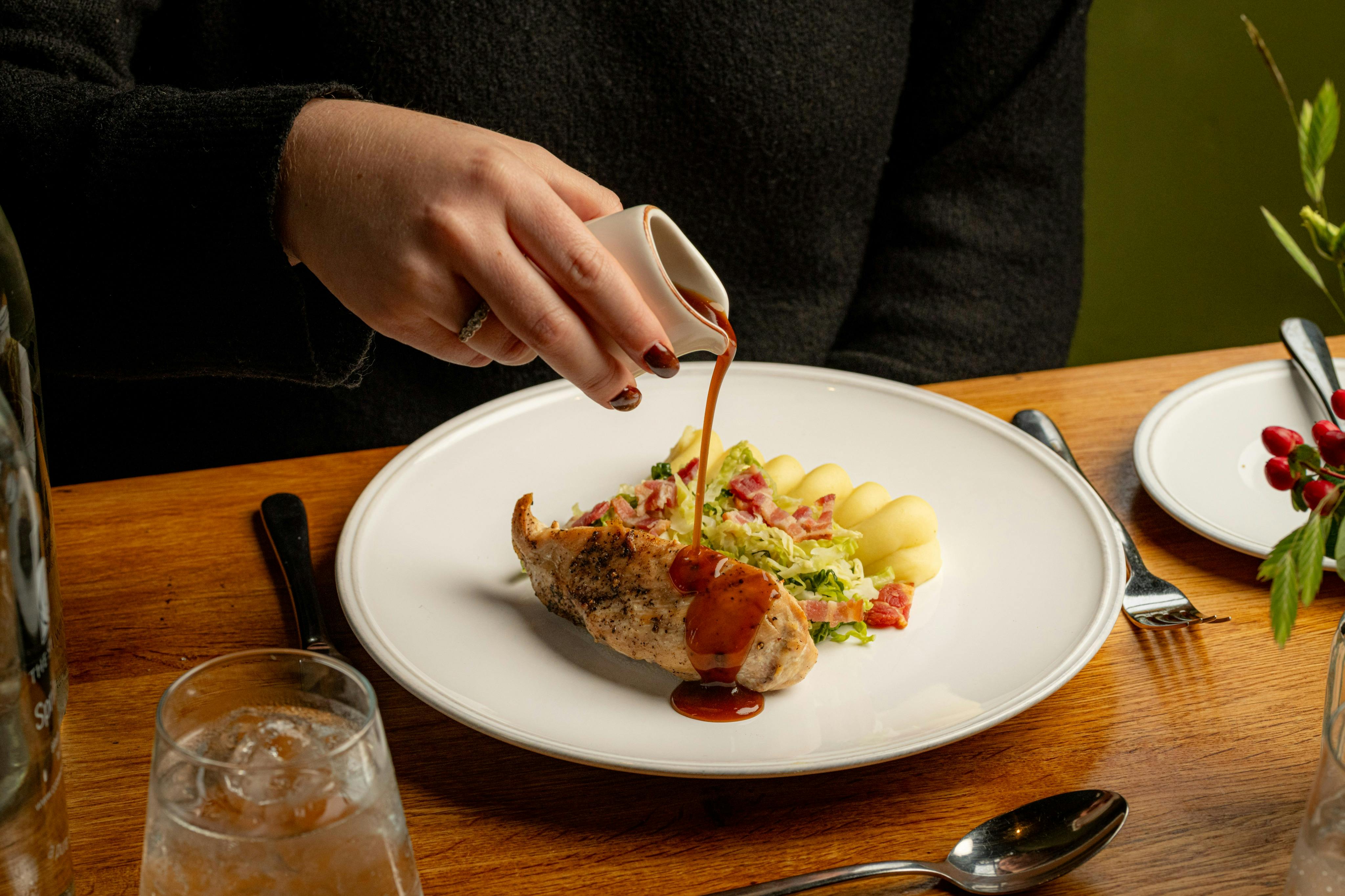 A person pours sauce from a small cup onto a plate of chicken, gnocchi, and vegetables on a wooden table.