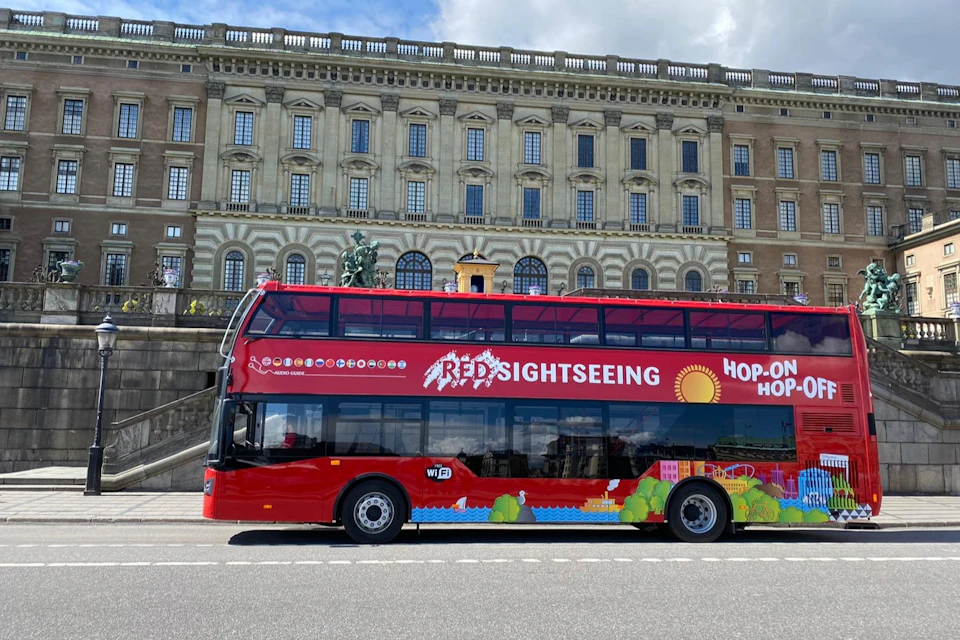 A red double-decker sightseeing bus parked in front of a large historic building under a partly cloudy sky.