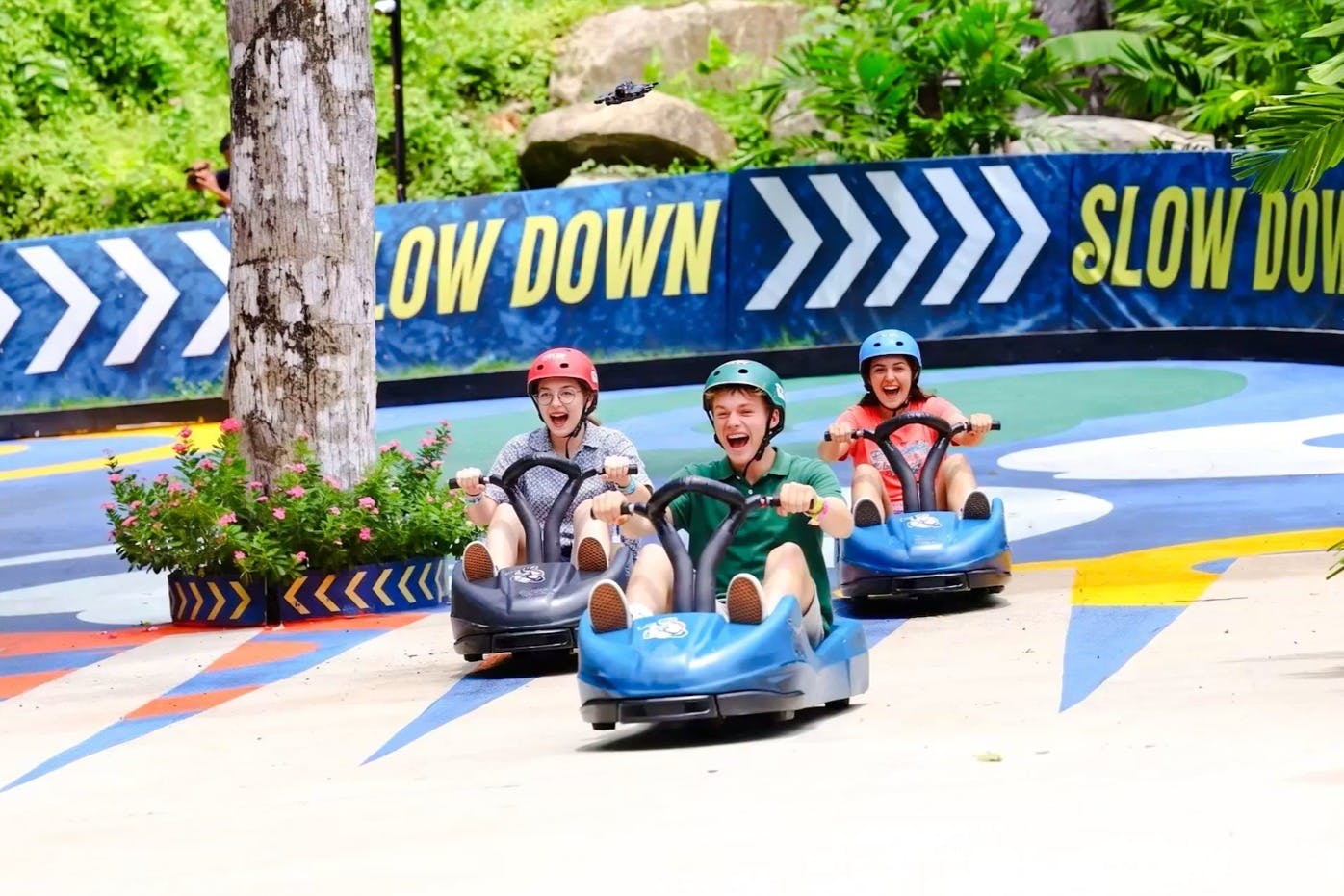 Trois personnes au volant de petits karts sur un circuit coloré, souriantes et portant des casques ; un panneau « Ralentissez » est visible à l'arrière-plan.