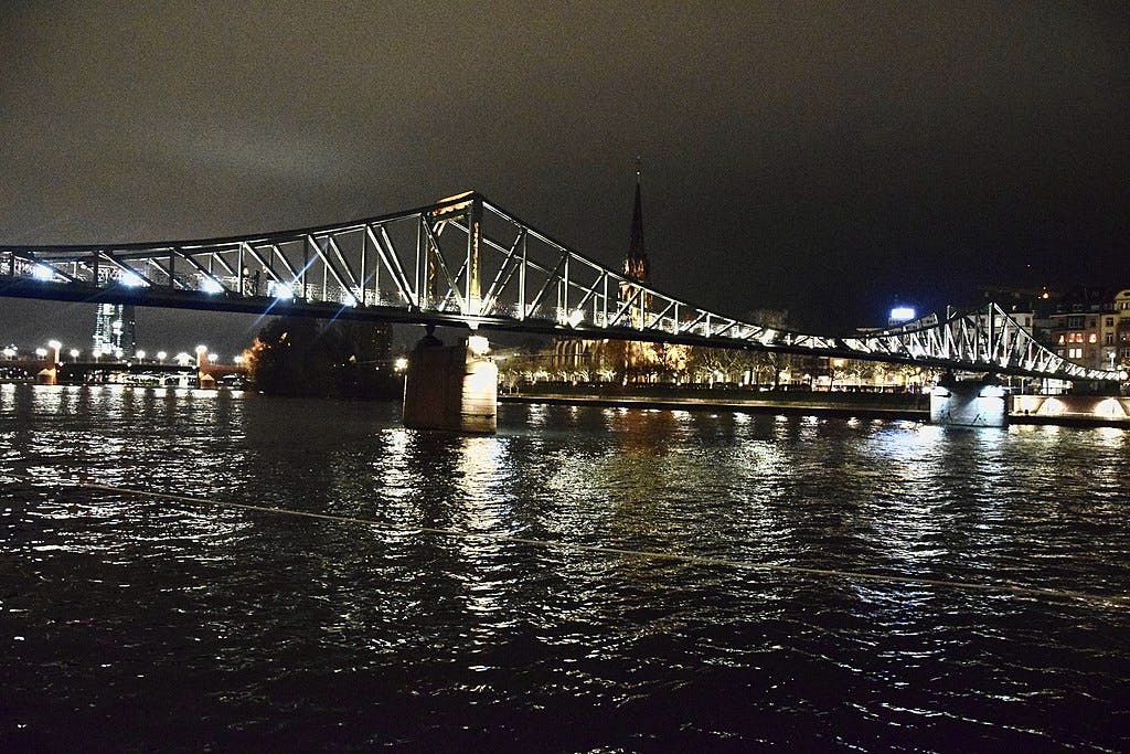 一座灯火通明的人行天桥在夜间横跨一条河，背景中可以看到城市建筑和教堂尖顶。