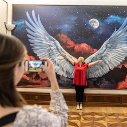 En person poserar med utsträckta armar framför en väggmålning av änglavingar när en annan person tar ett foto med en smartphone.
