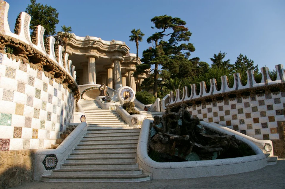 Ornate staircase leading to large columns, surrounded by mosaic walls and lush trees under a clear blue sky.