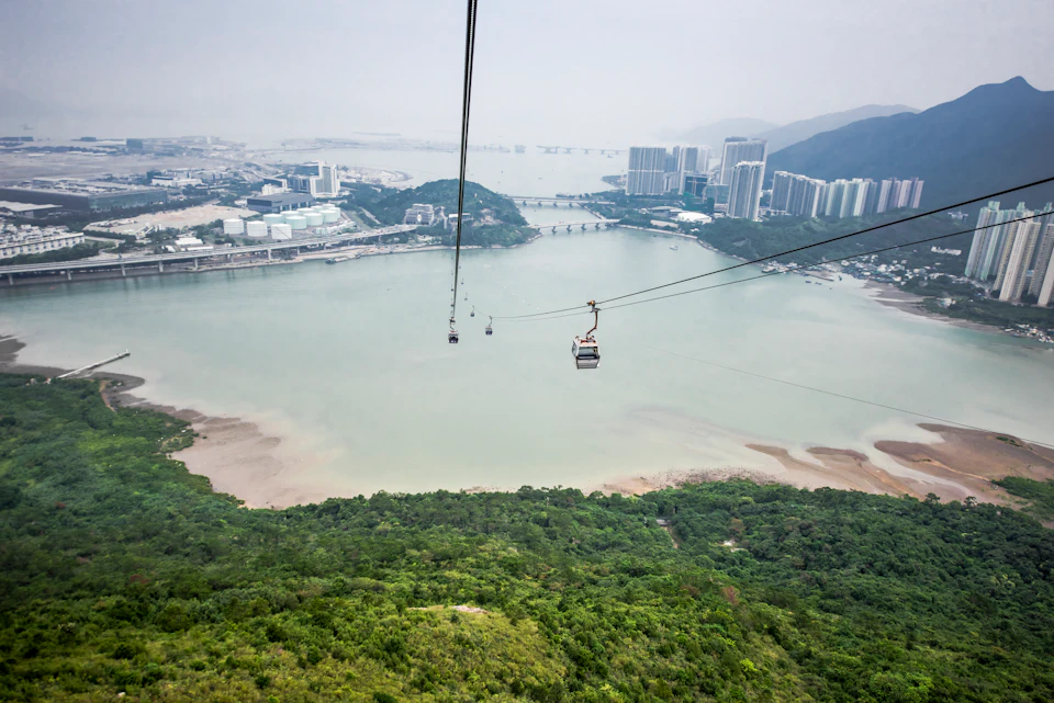 Suspended cable cars above a body of water with a cityscape in the background, set against green hills and a hazy sky.