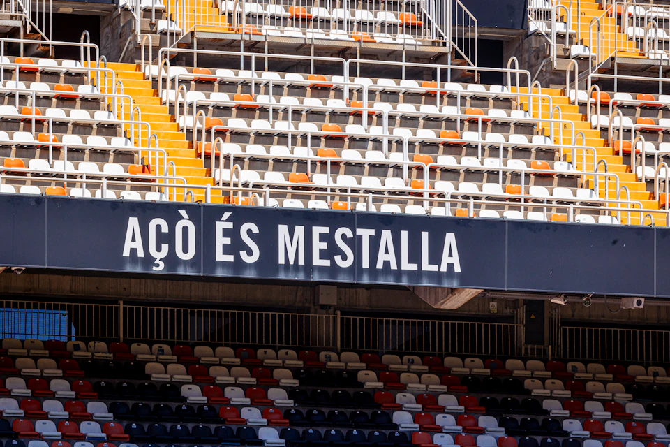 Empty stadium seats in shades of orange, red, and white, with a sign reading "ACÒ ÉS MESTALLA" in the foreground.