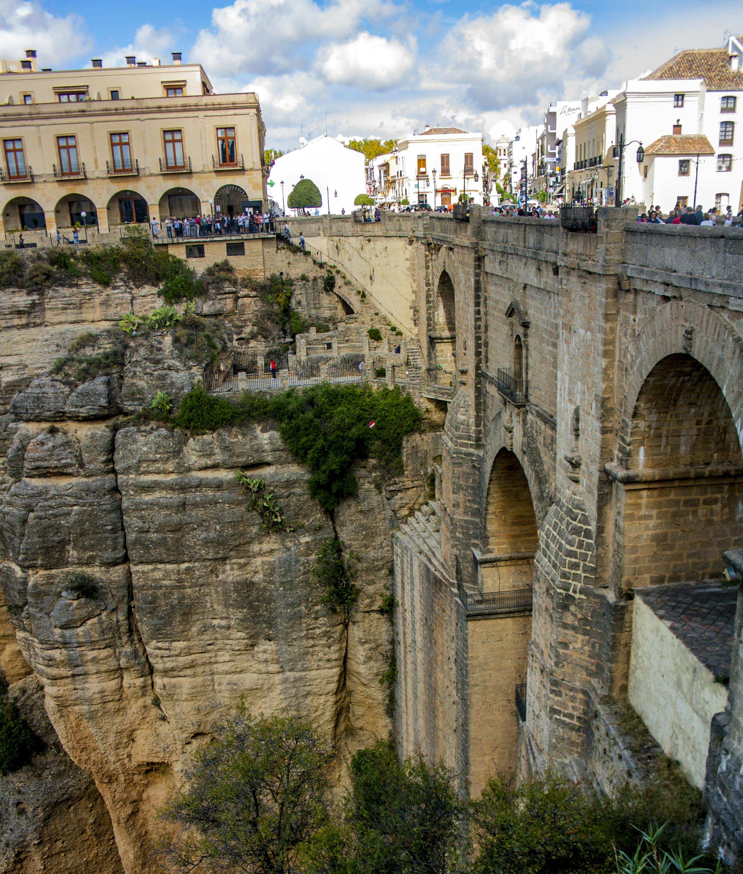 歴史的な石造りの橋は、深い岩の渓谷にまたがり、2つの町を建物と上から見ている人々で結んでいます。