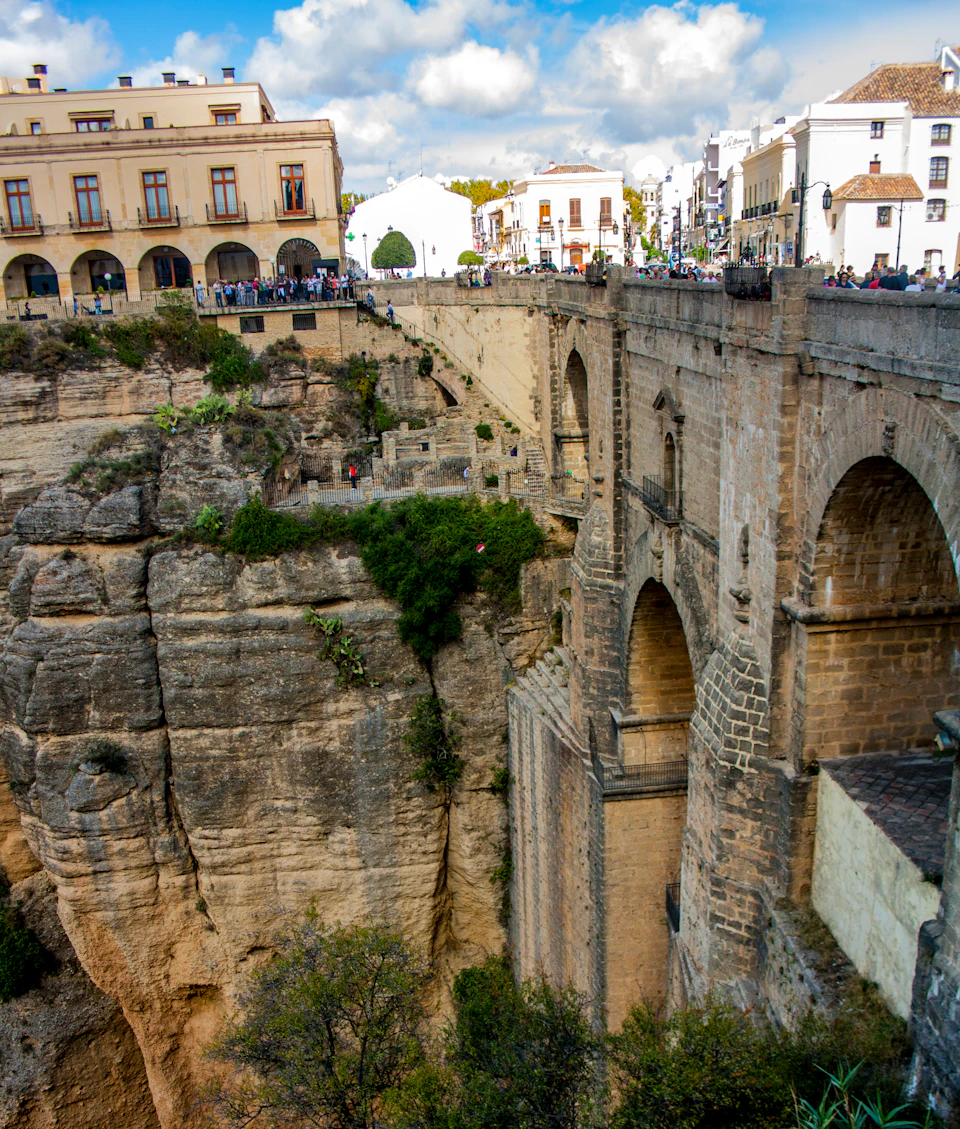 A historic stone bridge spans a deep rocky gorge, connecting two town areas with buildings and people observing from above.