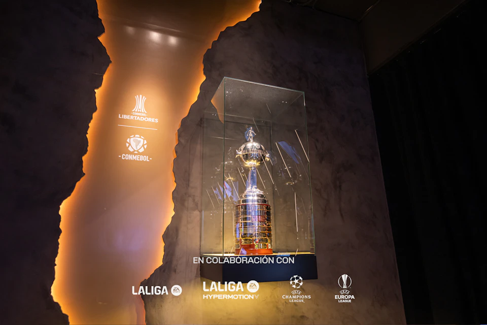 A silver trophy in a glass case, with various football league logos and the text "EN COLABORACIÓN CON" on a dark, textured background.