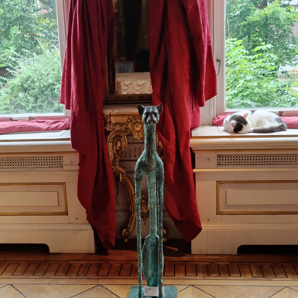 A tall, slender cat sculpture stands indoors in front of a large window with red curtains; a cat is napping on the windowsill.