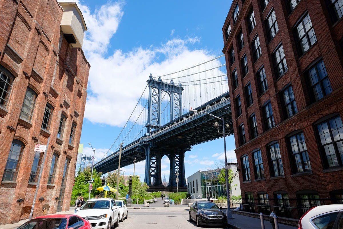 A tall blue suspension bridge viewed from a street with parked cars and brick buildings on both sides. Bright, sunny day.