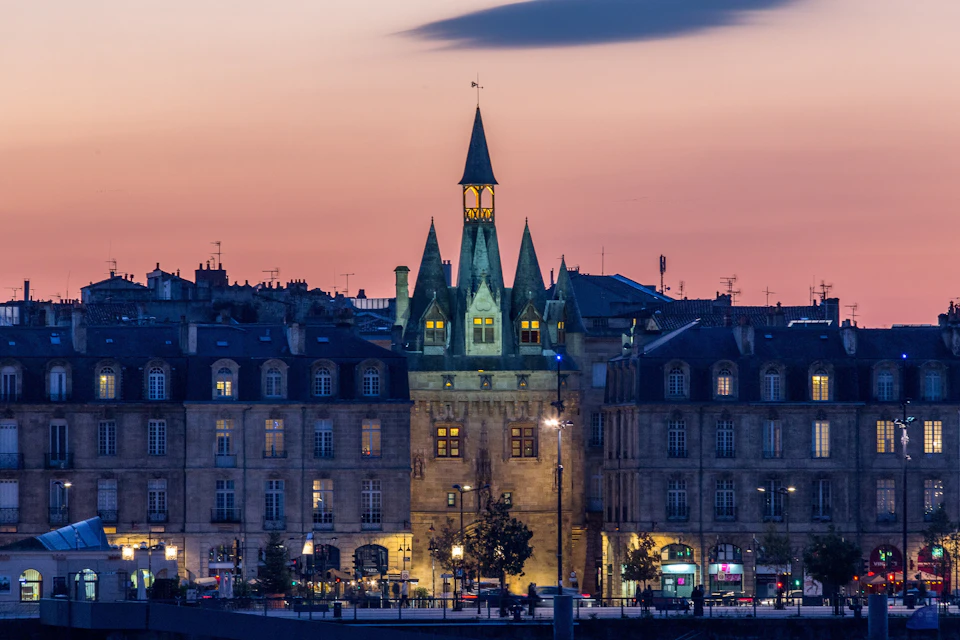 A historic building with pointed spires illuminated at dusk, surrounded by other buildings under a pink and blue sky.