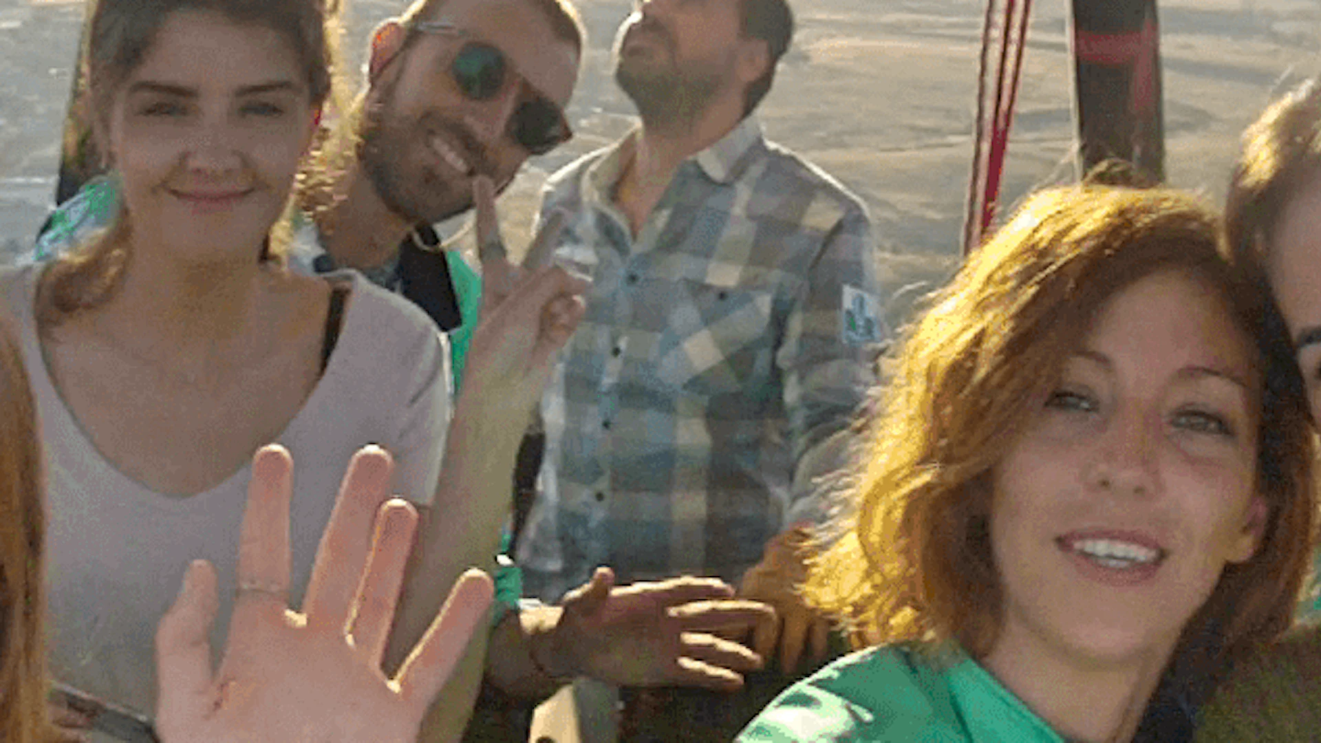 Five people smiling and waving inside a hot air balloon basket with a landscape visible in the background.
