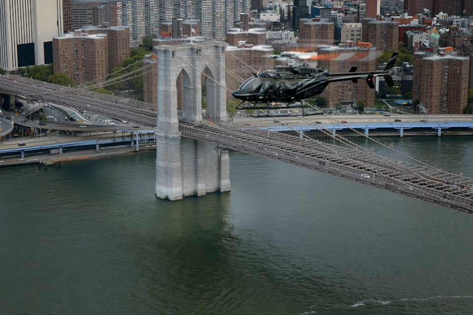 Helicopter flying near a stone tower of the Brooklyn Bridge over the East River, with city buildings in the background.