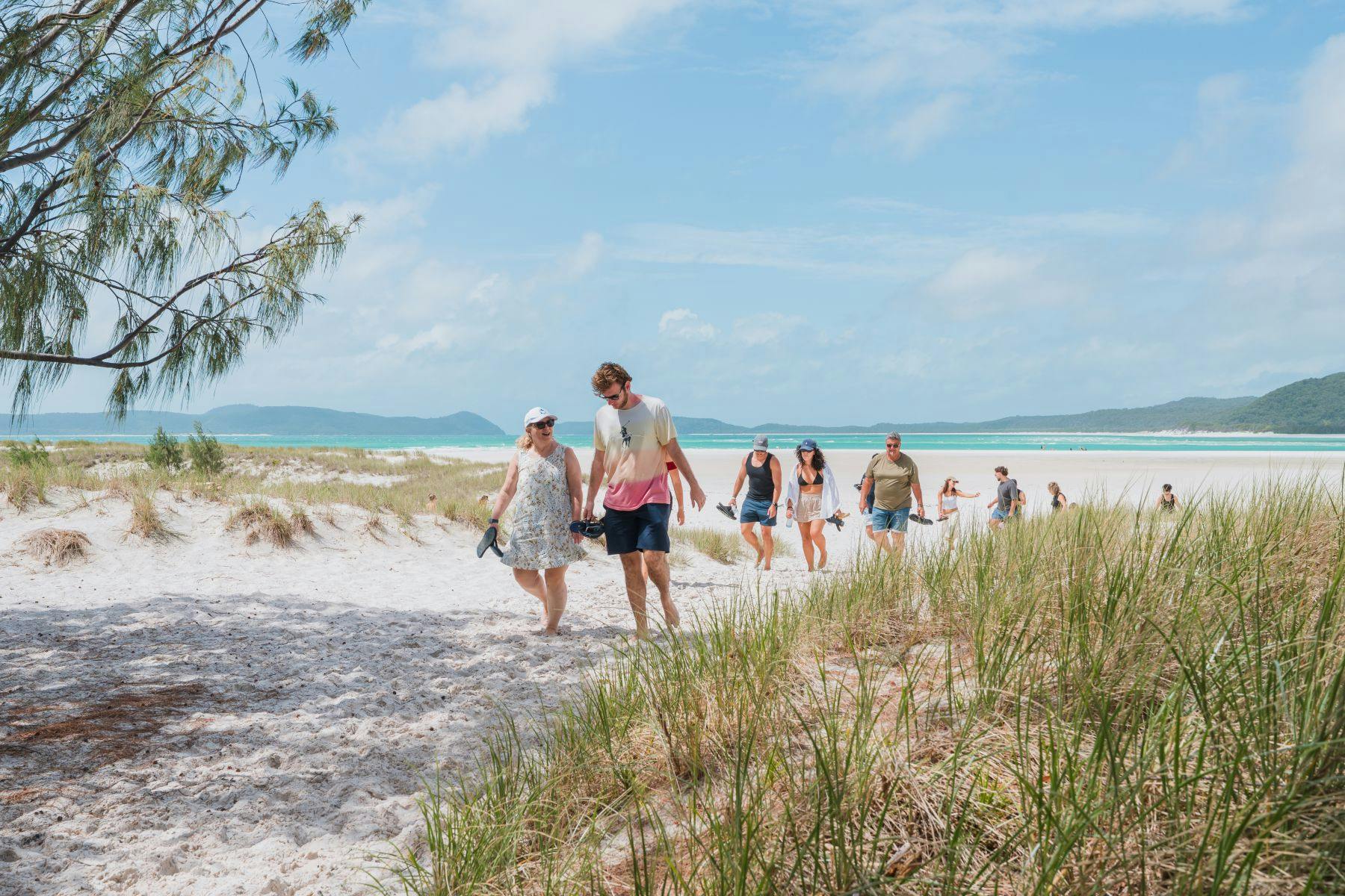 A group of people walking on a sandy beach path surrounded by grass and dunes under a partly cloudy sky near the ocean.