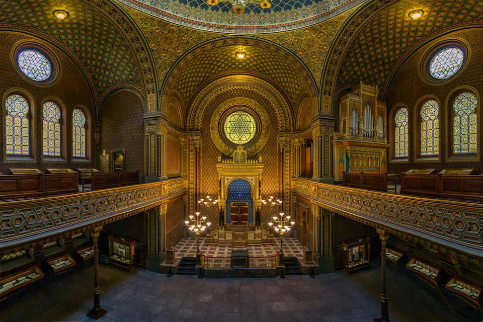 Ornate interior of a synagogue with an intricately decorated altar, symmetrical balconies, a stained glass window, and chandeliers.