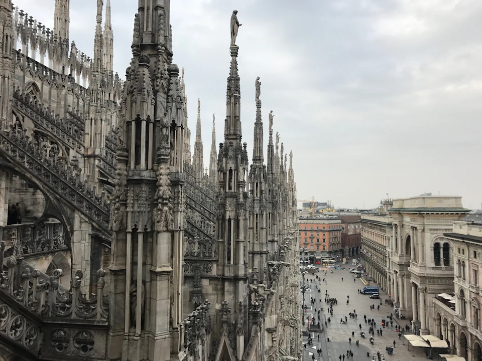 Ornate spires of a Gothic cathedral overlook a busy city square, with people walking and buildings in the background.