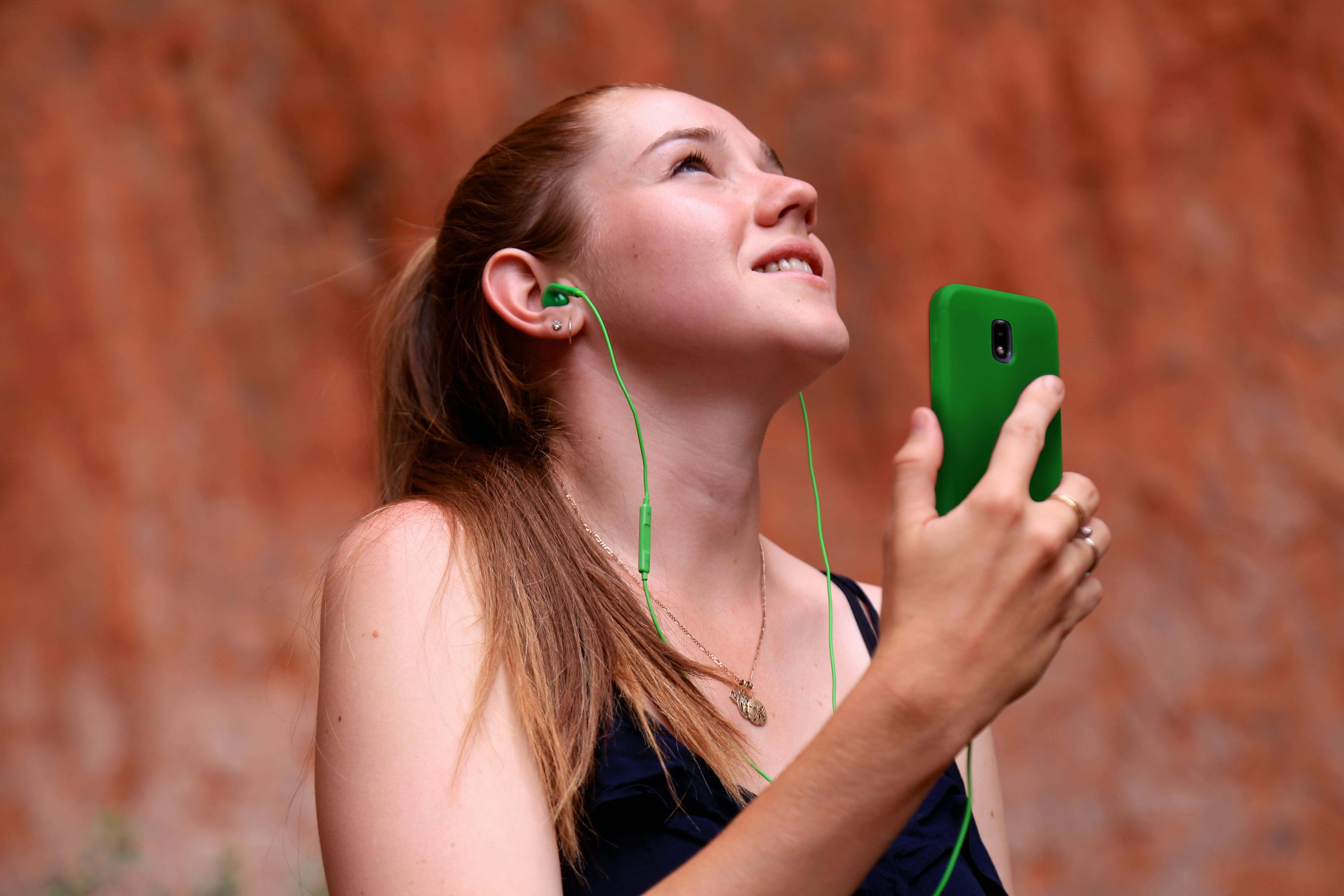 Una persona que lleva auriculares verdes y sostiene un teléfono verde mira hacia arriba, sonriendo, sobre un fondo marrón rojizo.