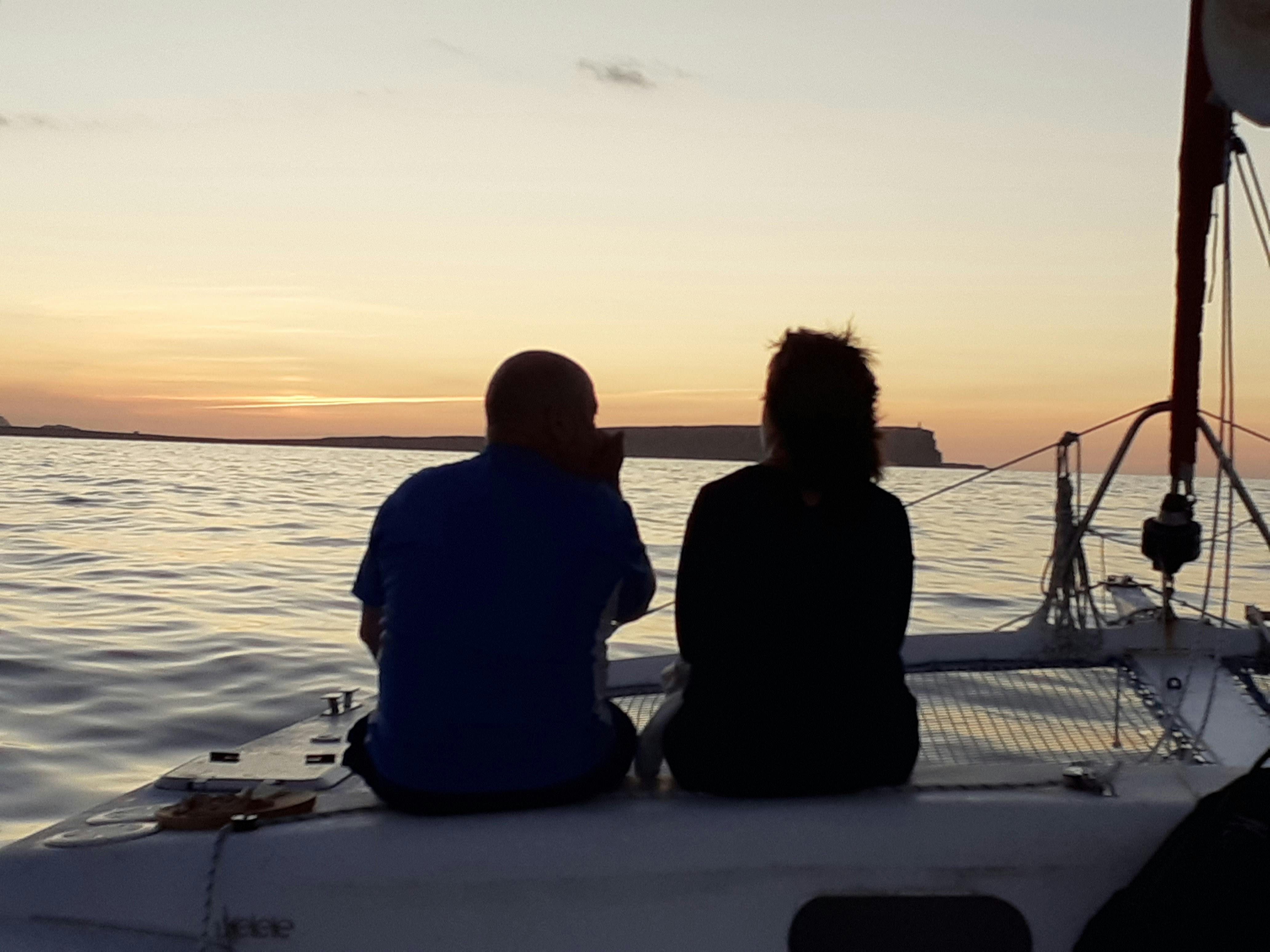 Two people sit on a boat at sunset, facing the water and distant land. The scene is calm with dim lighting.
