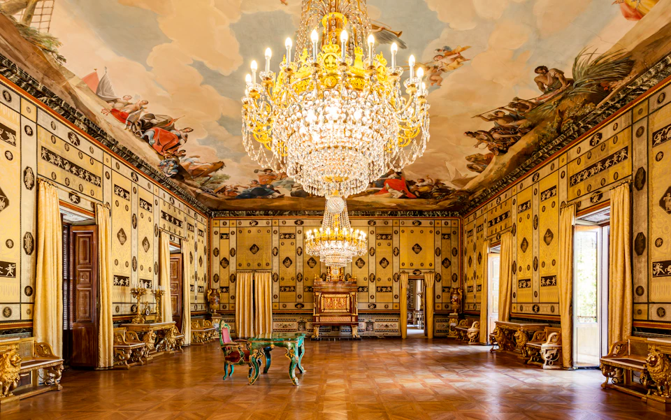 Ornate room with large chandeliers, murals on walls and ceiling, antique furniture, and parquet flooring.