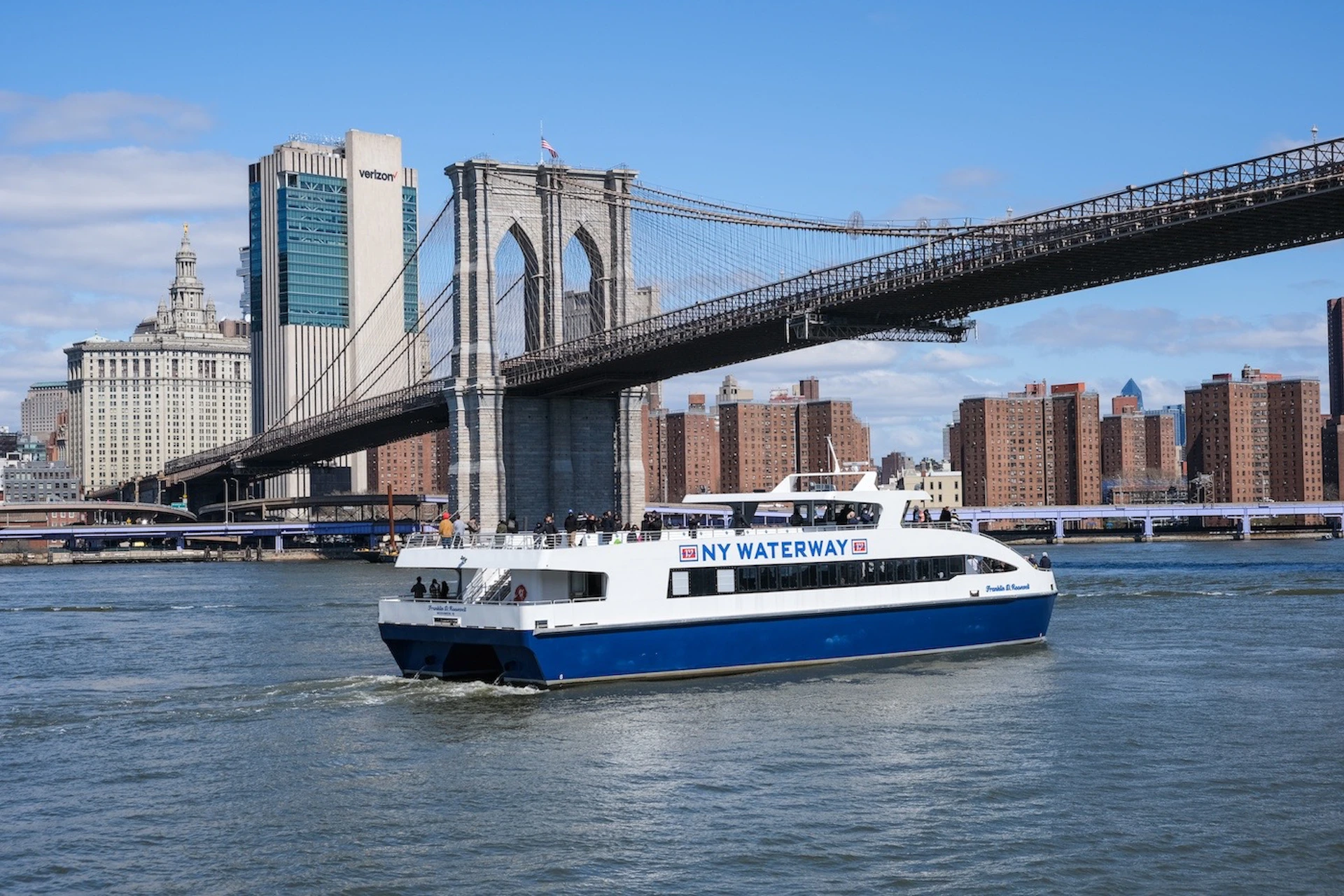 A NY Waterway ferry on the river with the Brooklyn Bridge and city buildings in the background under a clear sky.