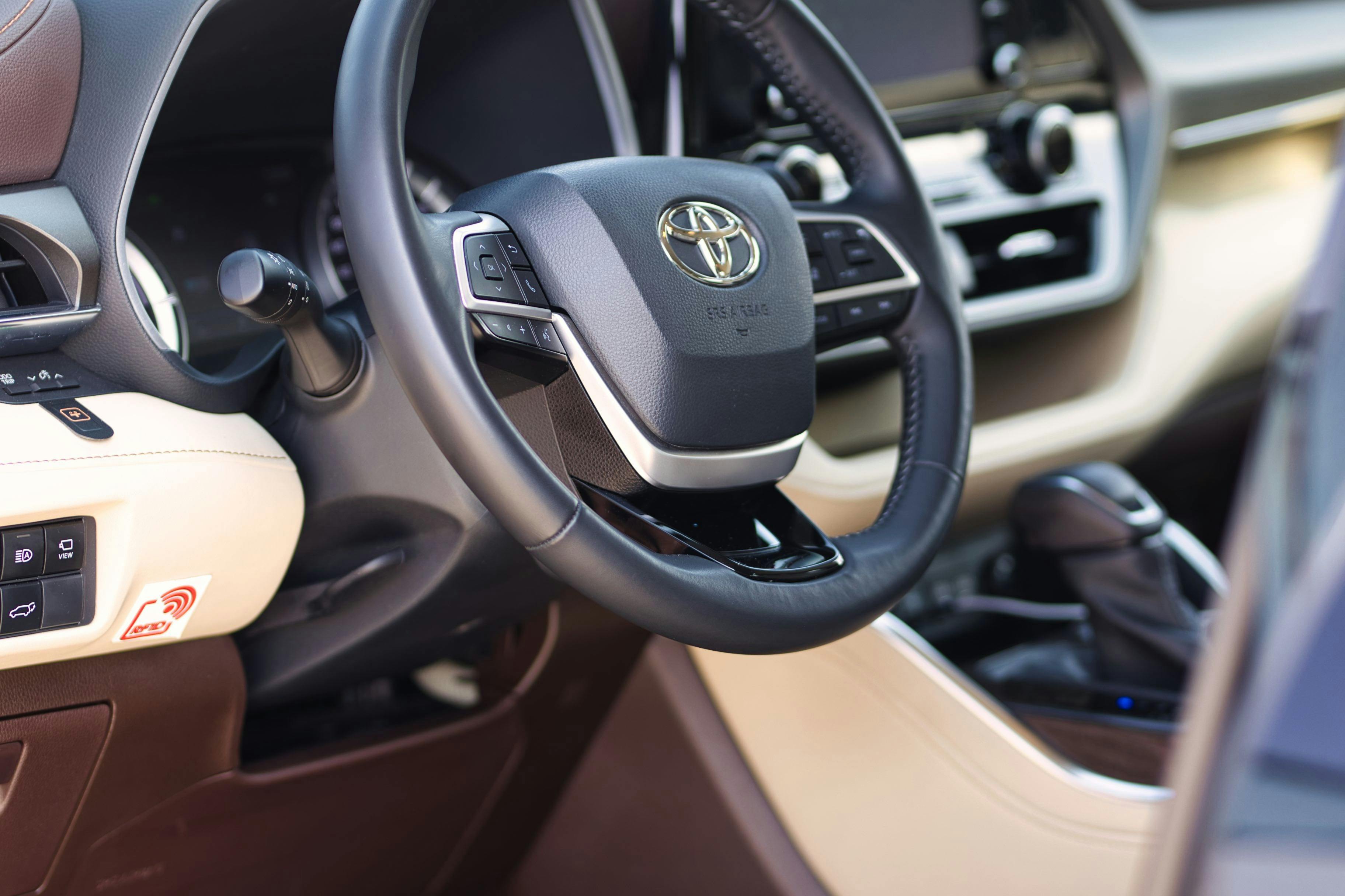 Närbild av en bils ratt med Toyota-logotypen, kontrollknappar och en del av den inre instrumentbrädan synlig.