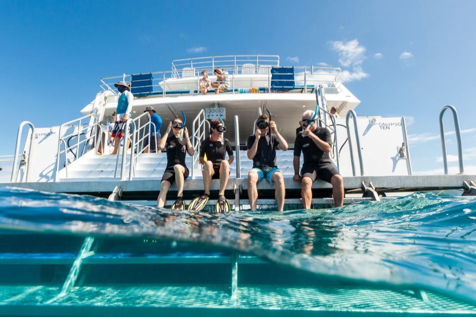 Quatre persones amb equip de busseig s'asseuen a la vora d'un vaixell, preparades per entrar a l'aigua. Altres estan a la coberta del vaixell de dalt.