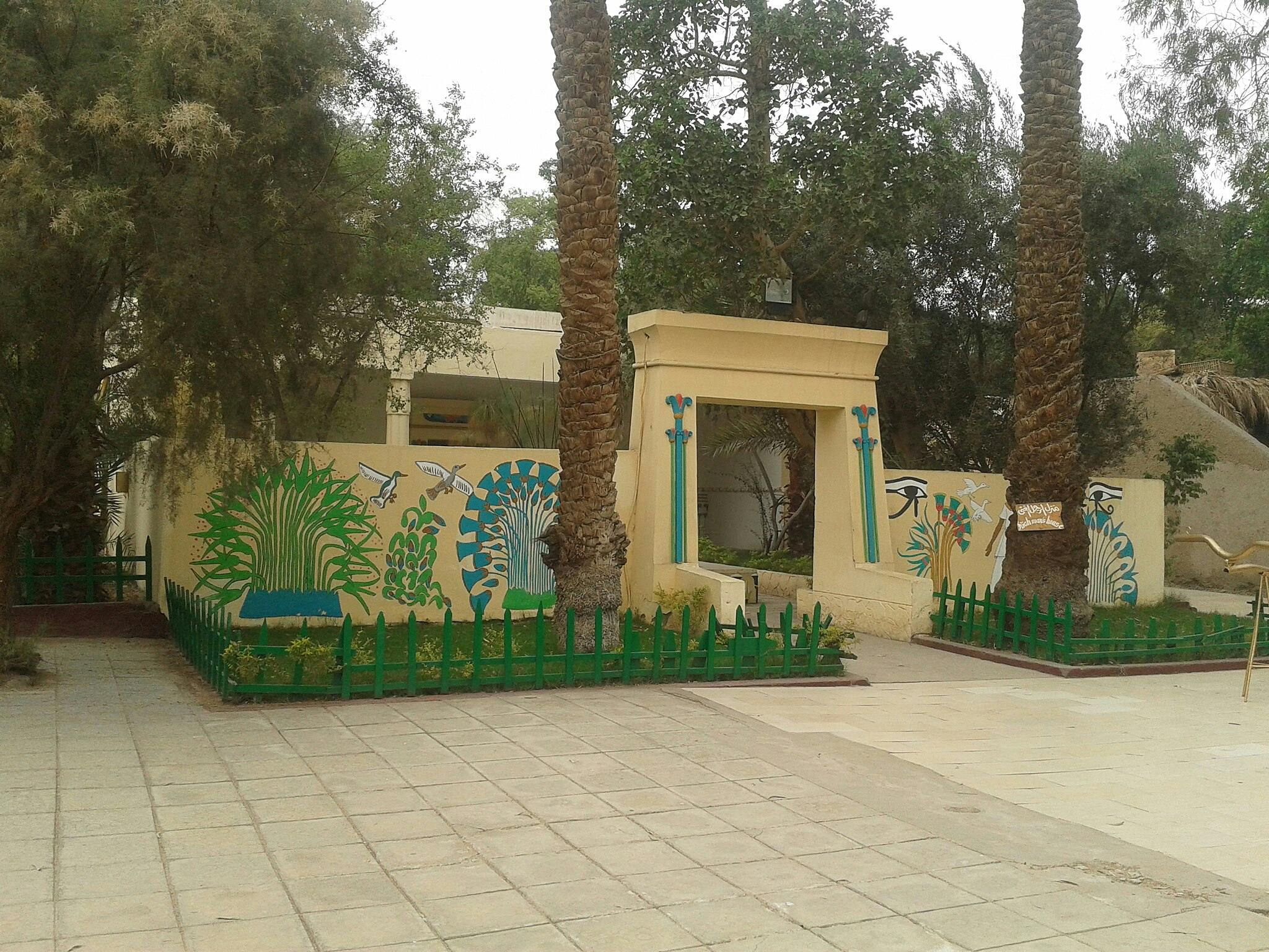 Вход в сад с фресками в египетском стиле и зелеными ограждениями, обрамленными высокими пальмами и кирпичными дорожками.