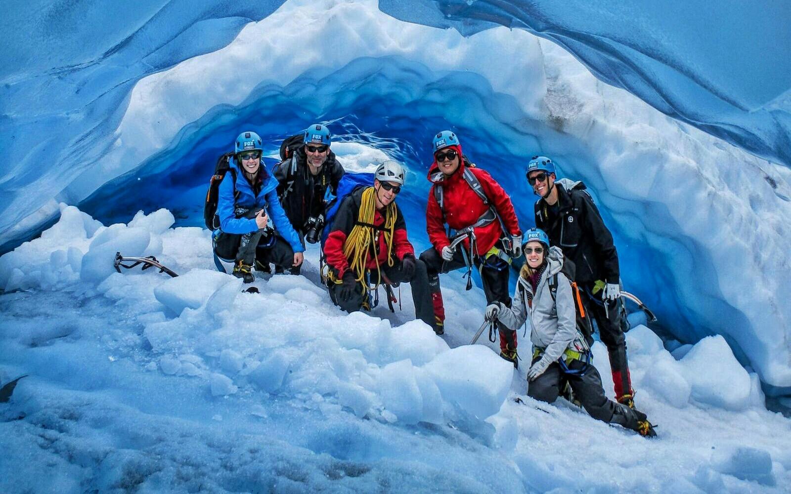 A group of six people wearing climbing gear and helmets posing inside a blue ice cave, surrounded by snow and ice.
