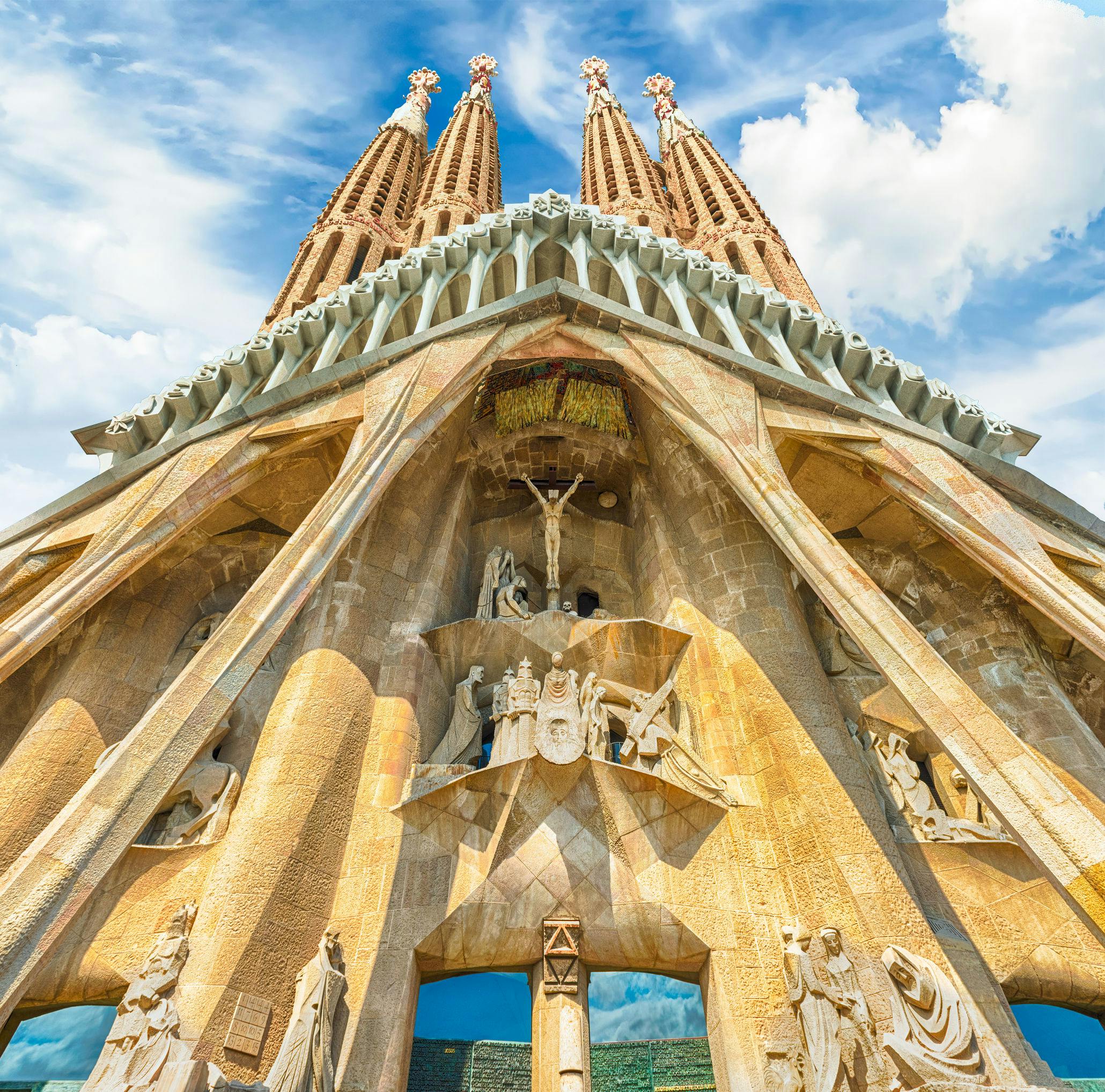 View from below of Sagrada Familia's intricate facade with detailed sculptures and tall spires against a cloudy blue sky.
