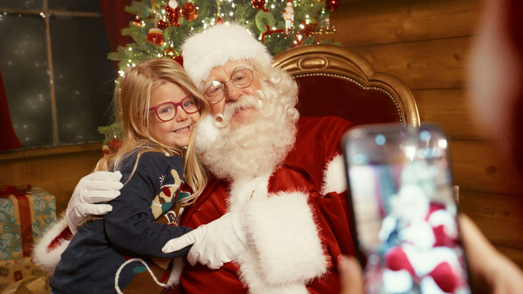 A girl wearing glasses hugs Santa Claus, sitting in front of a decorated Christmas tree with presents in the background.