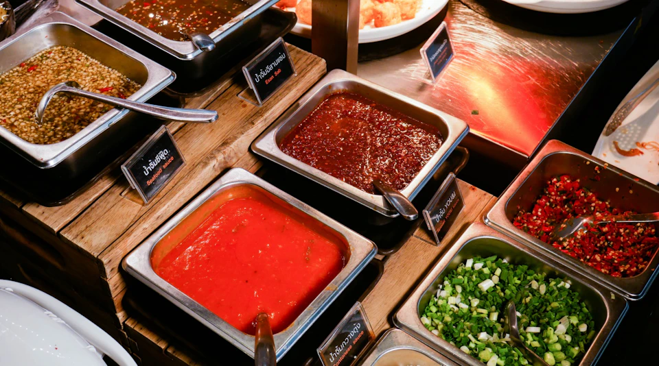 A selection of sauces and condiments in metal trays on a buffet counter, accompanied by small signs with labels.