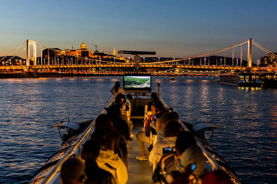 People seated on a boat at twilight, with a cityscape featuring illuminated buildings and bridges in the background.