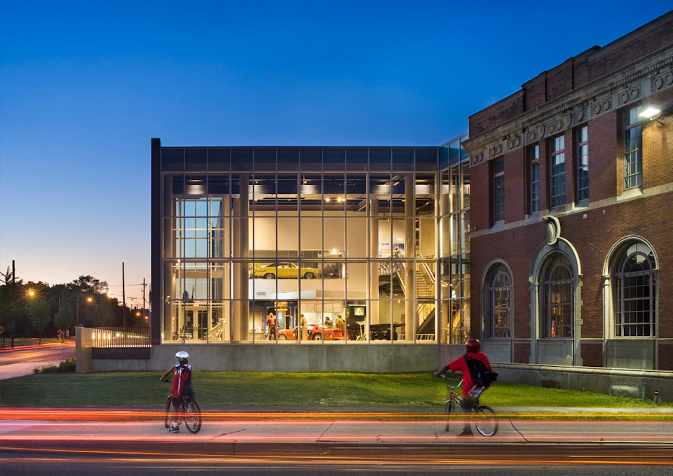 Two cyclists pass by a brightly lit, modern glass building next to a brick structure at dusk.