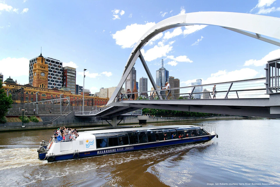 A modern bridge over a river, with a sightseeing boat passing underneath and a city skyline in the background.