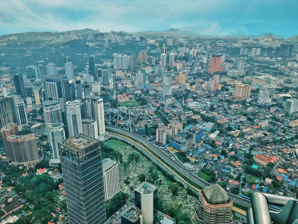 Aerial view of a city with numerous skyscrapers, buildings, a curving highway, and patches of greenery, set under a partly cloudy sky.