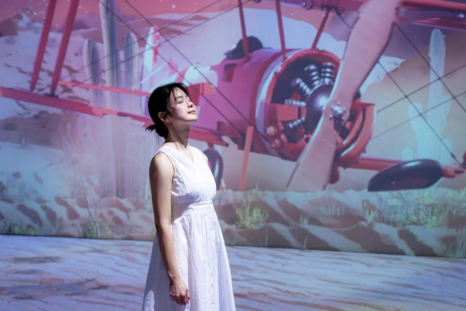 一位身穿白色连衣裙的女士闭着眼睛站在沙漠场景中红色双翼飞机的壁画前。