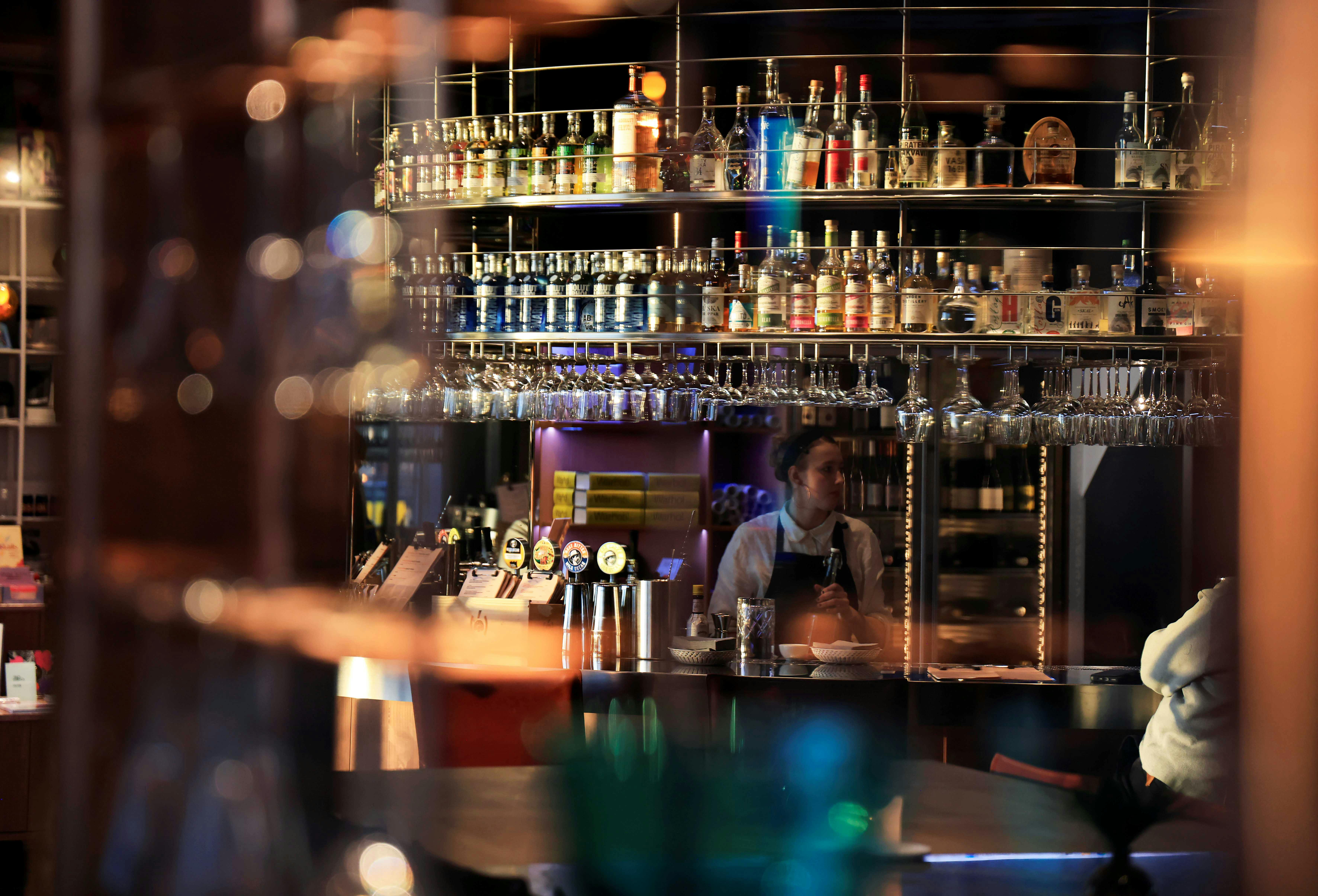 Un barman se tient derrière un comptoir dans un bar avec des étagères de bouteilles et de verres, reflétant une lumière chaude et atmosphérique.