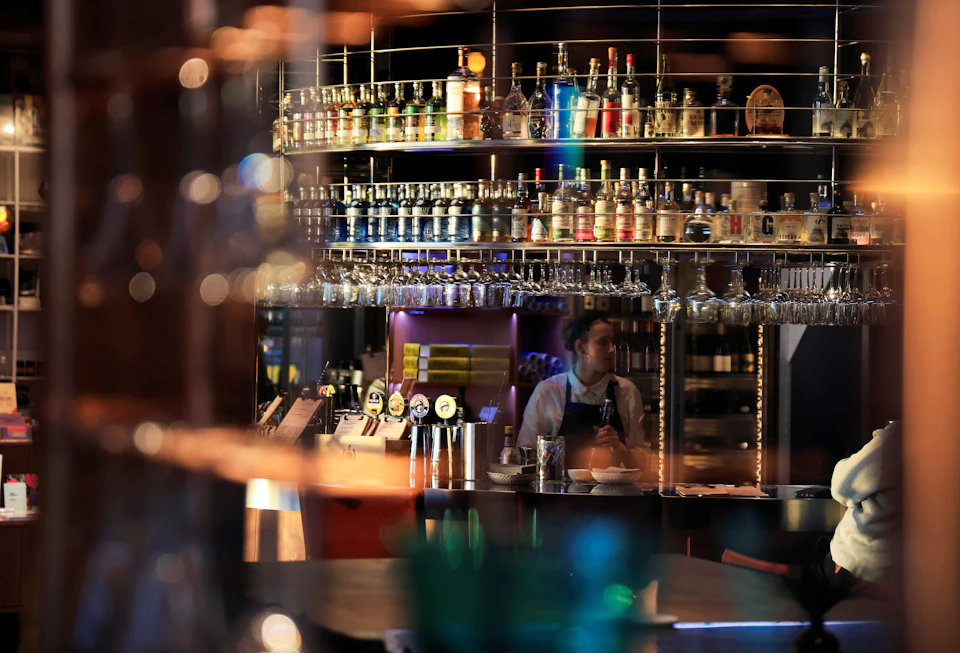 A bartender stands behind a counter in a bar with shelves of bottles and glasses, reflecting a warm, atmospheric light.