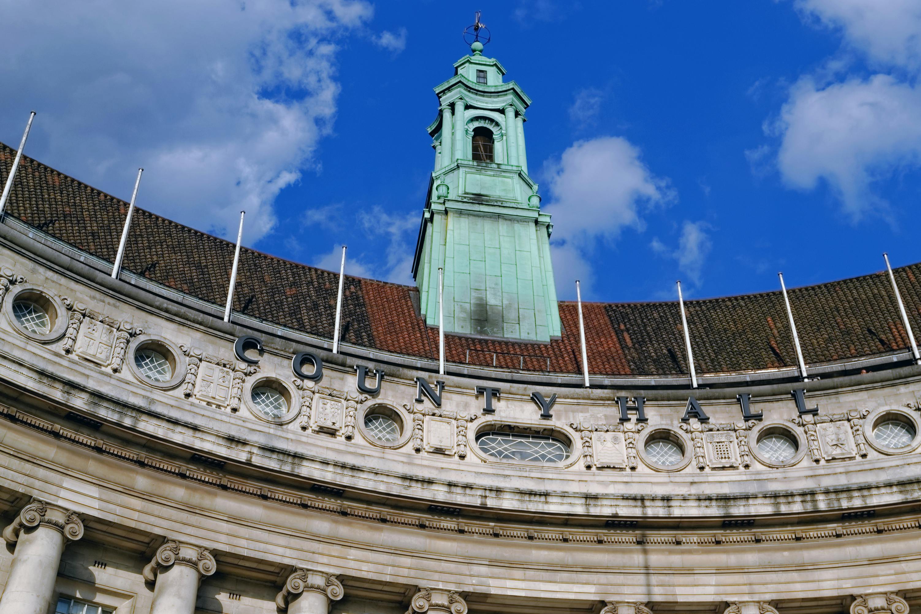 Facciata curva del County Hall con architettura ornata, finestre rotonde e una torre dell'orologio verde sotto un cielo blu.