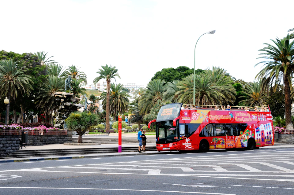 A red double-decker tour bus near palm trees in a park setting; two people stand by a bus stop.