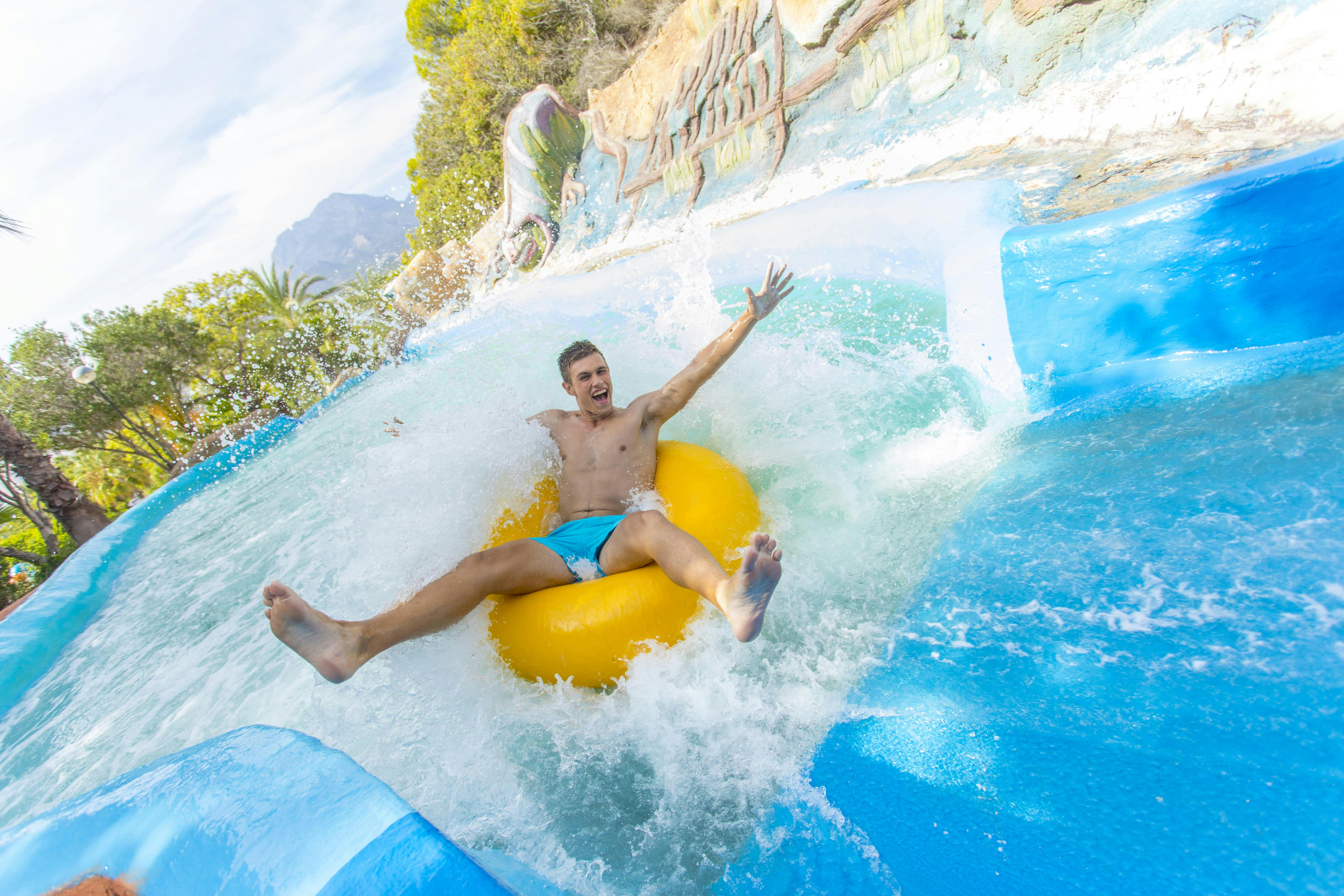 Man joyfully sliding down a water slide on a yellow tube, with water splashing around in a sunny outdoor setting.