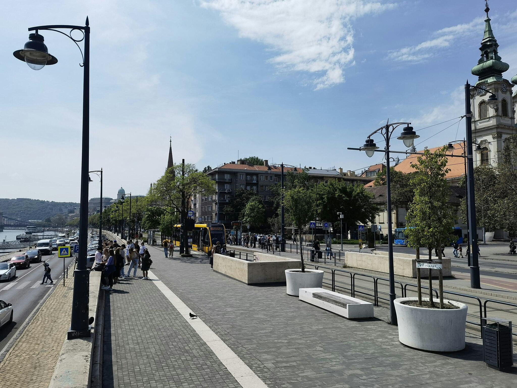 Θέα στον δρόμο με ανθρώπους να περιμένουν κοντά σε μια στάση του τραμ. Αυτοκίνητα και τραμ στο δρόμο, κτίρια και δέντρα στο παρασκήνιο. Φώτα στους δρόμους.