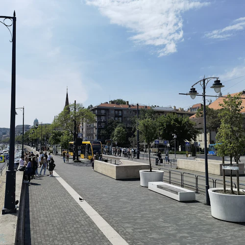 Gatuvy med människor som väntar nära en spårvagnshållplats. Bilar och spårvagnar på vägen, byggnader och träd i bakgrunden. Gatubelysning ovanför.