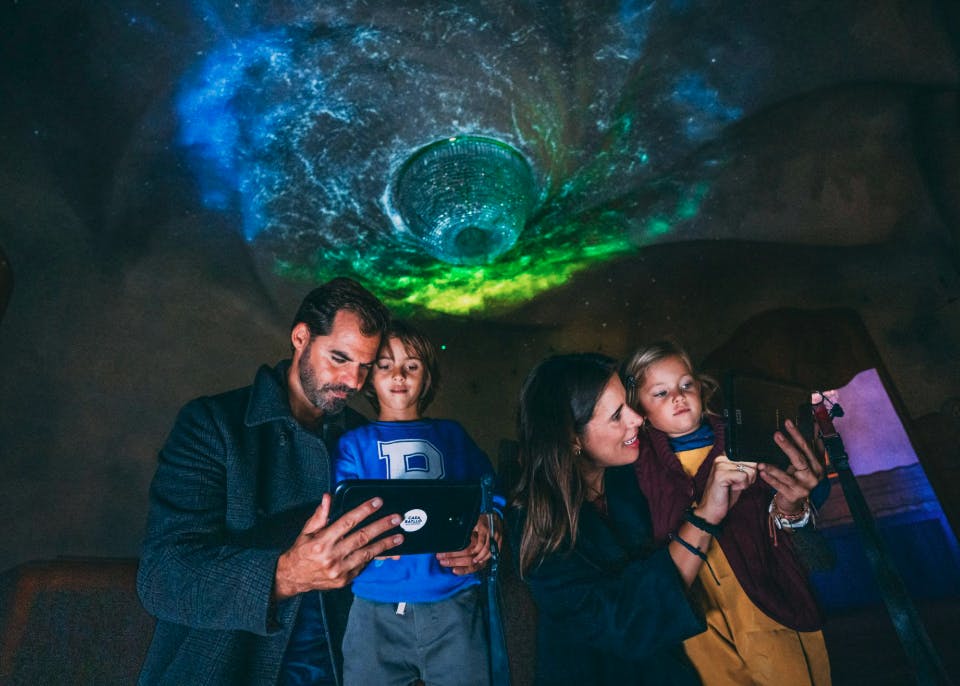 A family interacts with digital tablets under a ceiling projection of colorful lights in a dimly lit environment.
