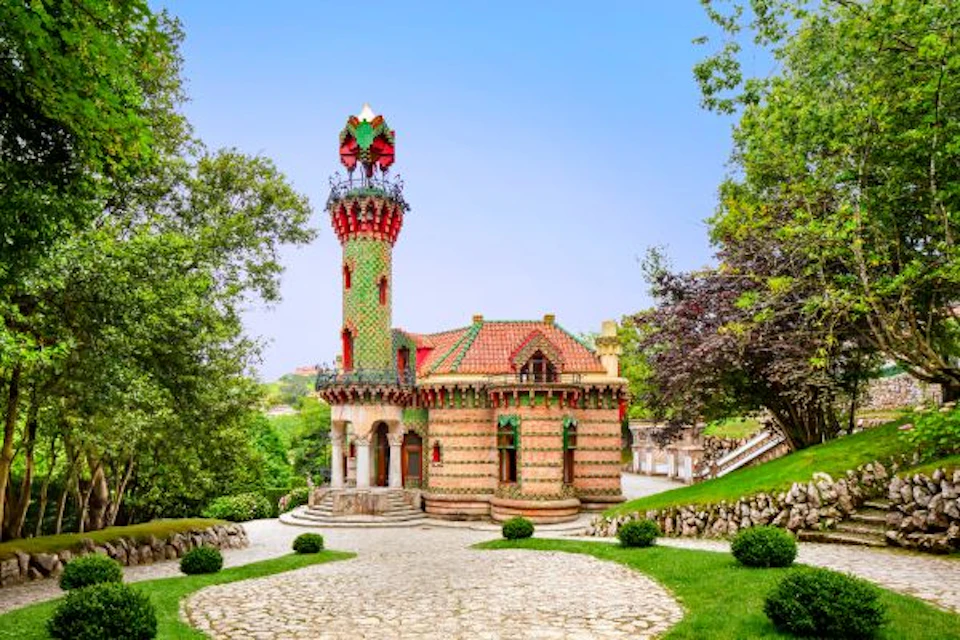 A colorful, ornate building with a tall tower, surrounded by greenery and a cobblestone path under a clear blue sky.