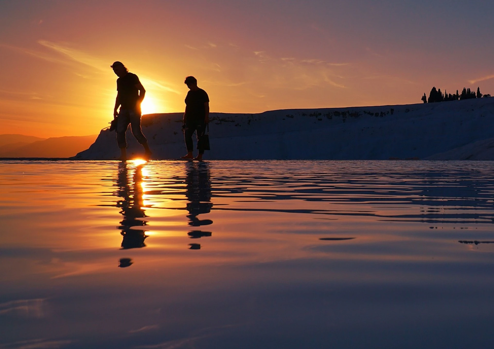 Two people walking through shallow water during sunset, with a silhouetted landscape in the background.