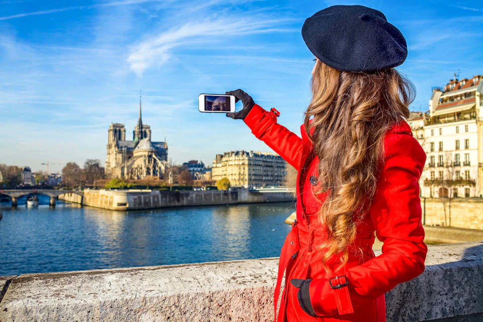 Person in a red coat and black beret taking a photo with a smartphone, capturing a riverside landmark under a blue sky.