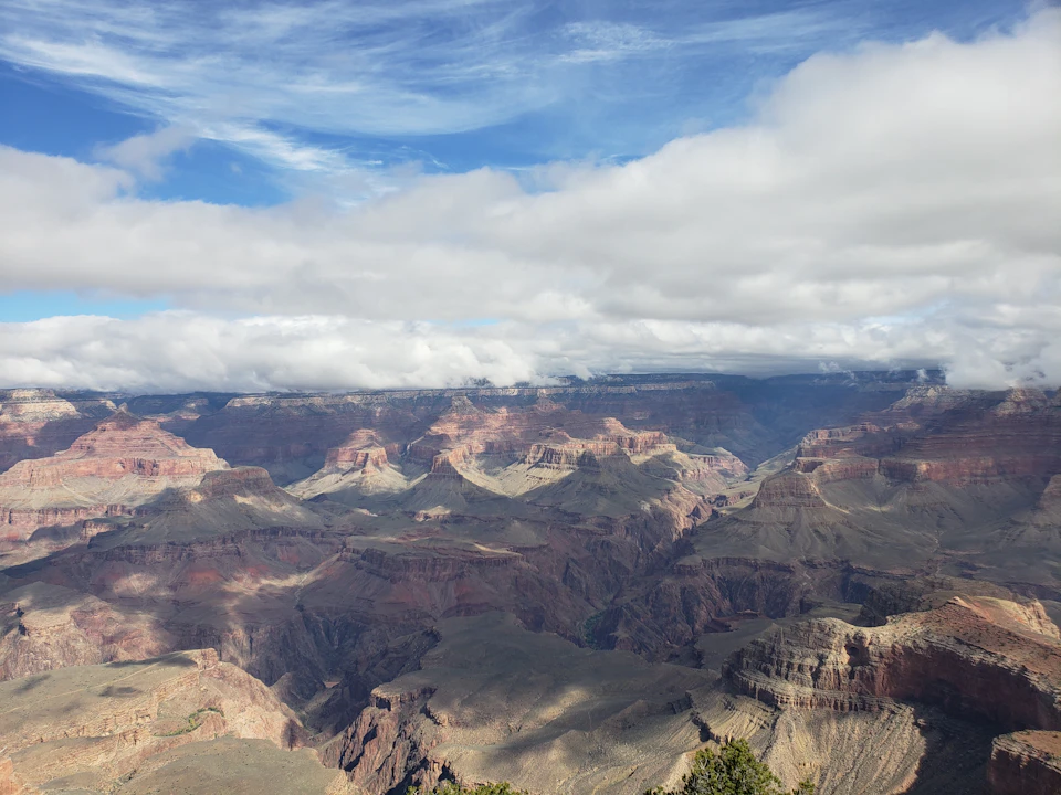 Clouds hover over the panoramic view of the Grand Canyon, showcasing layered rock formations under a partly cloudy sky.