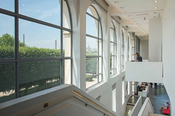 Moderní interiér s velkými klenutými okny s výhledem do parku a na památky, s osobou sedící na horním balkoně.