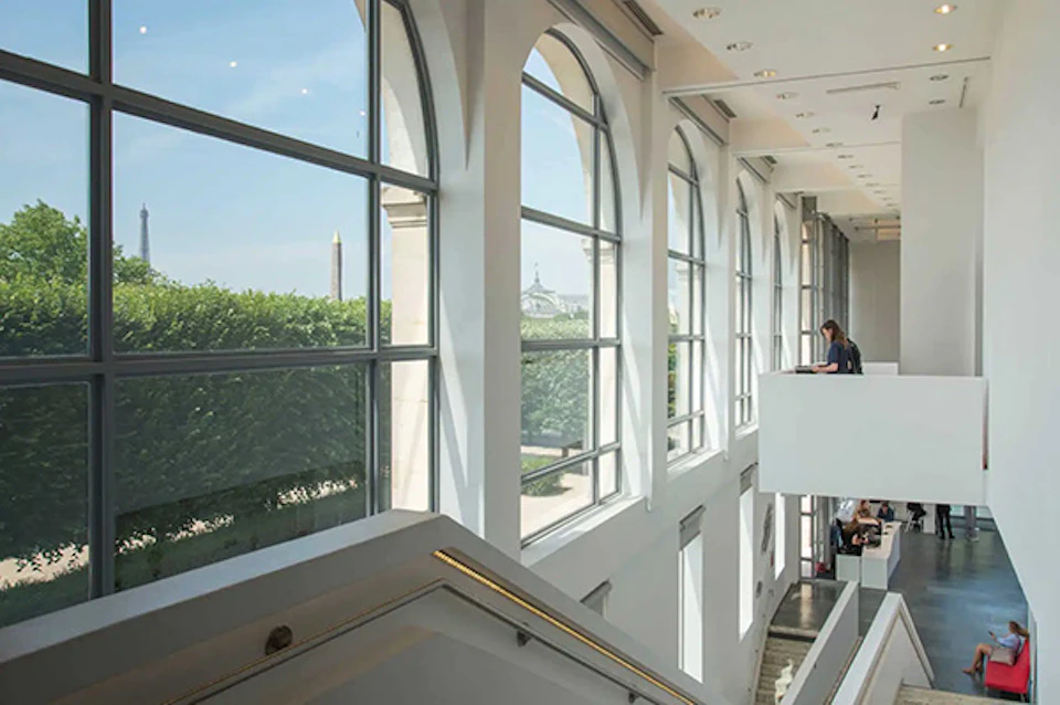 Modern interior with large arched windows overlooking a park and monuments, with a person sitting on an upper balcony.